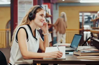 Woman with headphones sits at a library table, writing in a notebook with a laptop nearby.