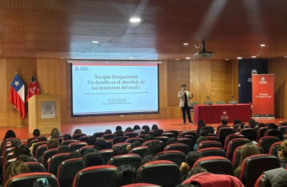 Lecture in a university auditorium with a presenter at a podium and a large screen showing the slide “Terapia Ocupacional: Un desafío en el abordaje de los trastornos del sueño.”
