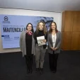 Three women pose for a photo in front of a university event sign, one holding a booklet.