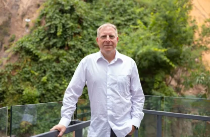Man in a white shirt leaning on a glass balcony railing with lush green plants behind him, smiling slightly.