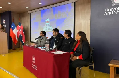 Four panelists seated at a red-draped table on a stage during a Universidad Andrés Bello event, with flags and a projection screen behind them.