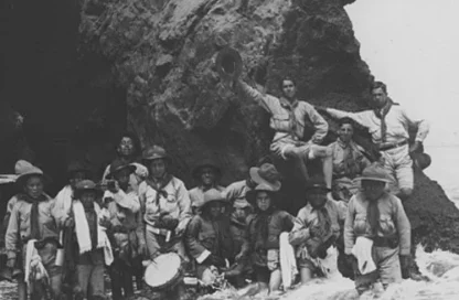 Black-and-white photo of a large group of scouts and adults posing on a rocky shoreline, some standing, others seated, with waves nearby.