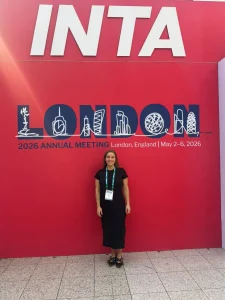 Woman in a black dress standing in front of a red INTA London 2026 banner and conference badge visible around her neck