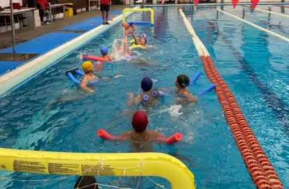 Children in a swimming pool during a lesson, using float noodles and kickboards as they splash and play near the lanes.