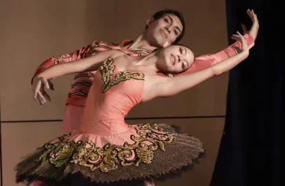 Two ballerinas in peach costumes with gold embroidery perform a duet on stage, one dancer leaning back into the other's support in a graceful pose.