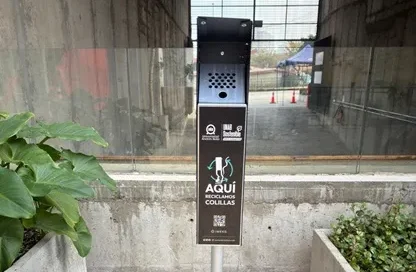 Tall black public waste/recycling station mounted on a post in an outdoor walkway, with a QR code on the front panel and a perforated vent area at the top.