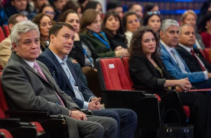 Audience at a conference: two men in the foreground in business attire, red seats behind them.