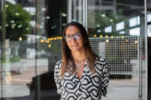 Smiling woman with glasses in a black-and-white zebra-print blouse stands outside glass doors at a modern building.