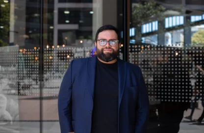 Man with dark hair and beard wearing glasses and a navy blazer stands in front of a glass storefront.