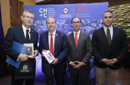 Four men in suits pose for a formal photo in front of a blue event backdrop, with two holding books or folders.