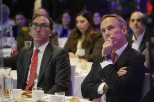 Two men in suits seated at a round banquet table, one with a red tie and glasses and the other resting his chin on his hand, as other guests sit in the blurred background with plates and drinks on the table.