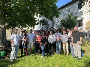 Group of people standing closely together outdoors under green trees, posing for a photo in a sunny setting.