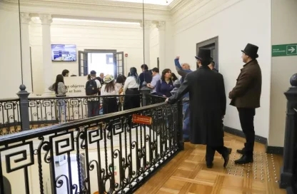 Group of visitors in a bright museum hallway, some in period costumes with top hats, looking at exhibits near a railing.