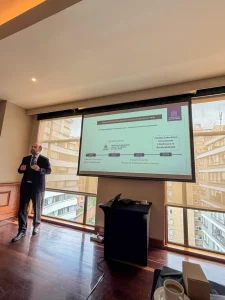 Man in a suit giving a presentation in a conference room with a projected slide and large windows overlooking buildings outside.