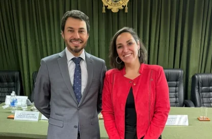 Two professionals pose for a photo in a conference room; man in a gray suit and tie, woman in a red jacket smiling at the camera.
