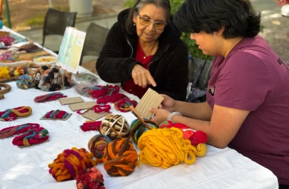 Two women at an outdoor market table examining handmade felt and yarn crafts, with bright skeins and woven items laid out.